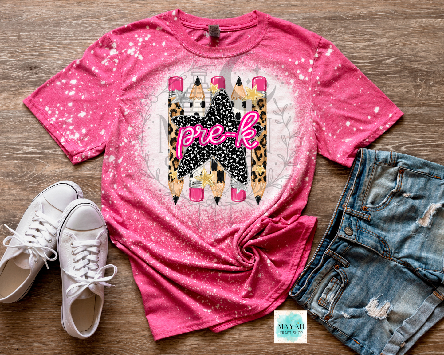 A pink short-sleeve graphic T-shirt laid flat on a wood surface, styled with white sneakers and distressed denim shorts. The shirt has a splatter/glitter finish and a large central graphic featuring the word 'pre-k' in pink script over a castle and pencils.