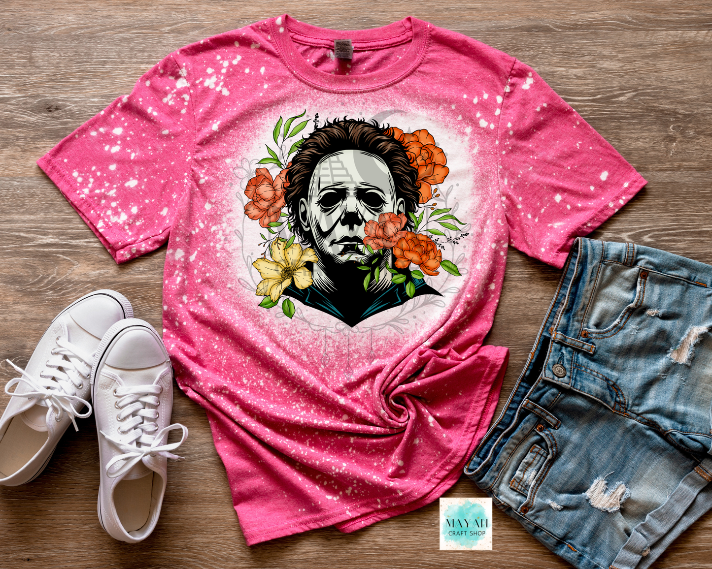 Pink t-shirt with a graphic design of a face surrounded by flowers, paired with white sneakers and denim shorts on a wooden surface.