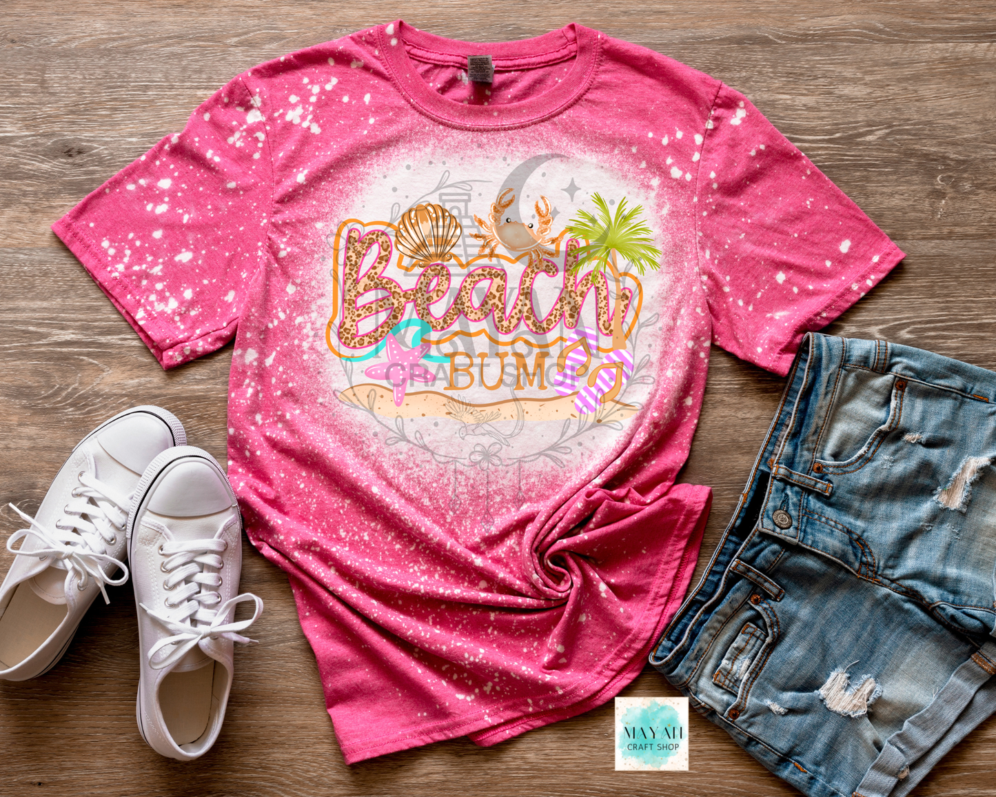 Pink 'Beach Bum' t-shirt with sunglasses, palm tree, and seashell graphic on a wooden surface with white sneakers and denim shorts.