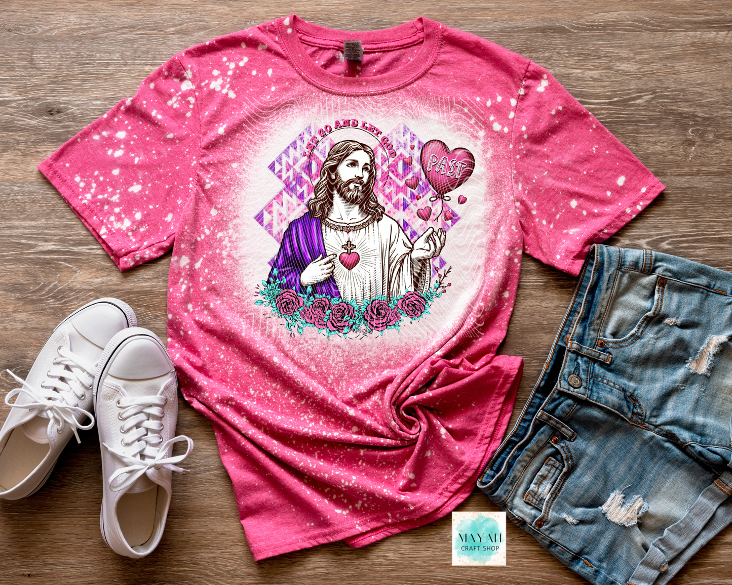 Pink t-shirt with a graphic design of a religious figure, surrounded by hearts and flowers, on a wooden surface with white sneakers and denim shorts.