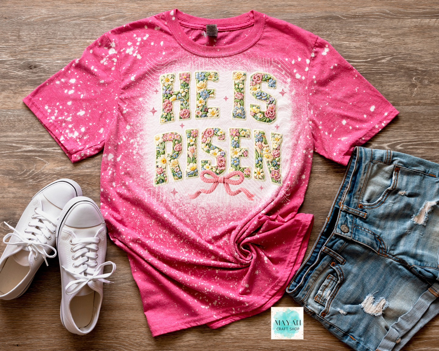 Pink t-shirt with 'He is Risen' text, white sneakers, and denim shorts on a wooden surface.