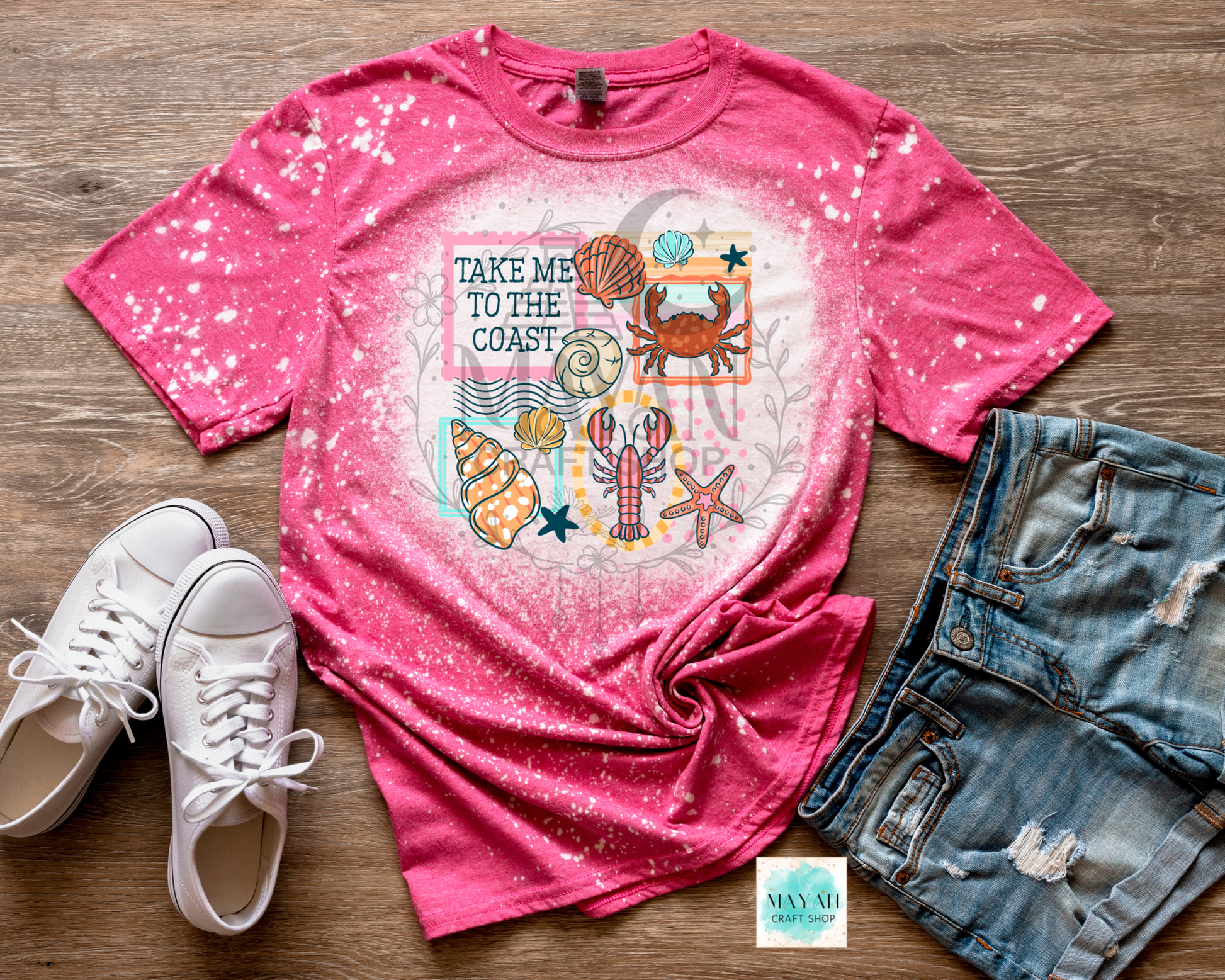 Pink t-shirt with coastal design, white sneakers, and denim shorts on a wooden surface
