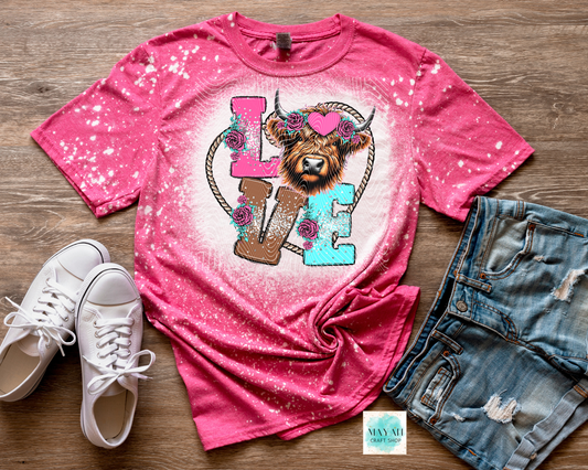 Pink t-shirt with a graphic design of a cow head, flowers, and 'LOVE' text on a wooden surface with white sneakers and denim shorts.