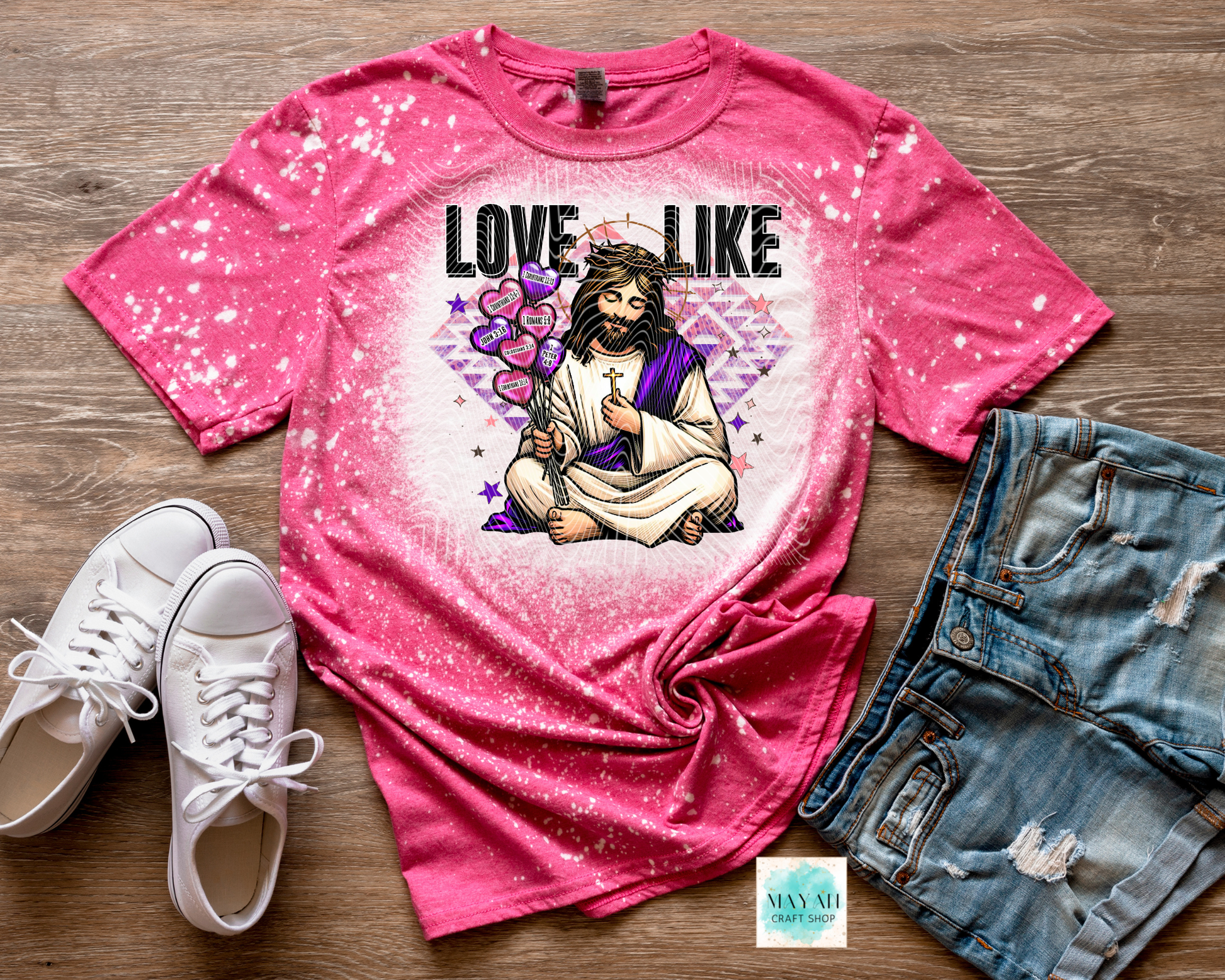 Pink t-shirt with graphic design of a person in a meditative pose, surrounded by hearts and text 'LOVE LIKE', paired with white sneakers and denim shorts on a wooden surface.
