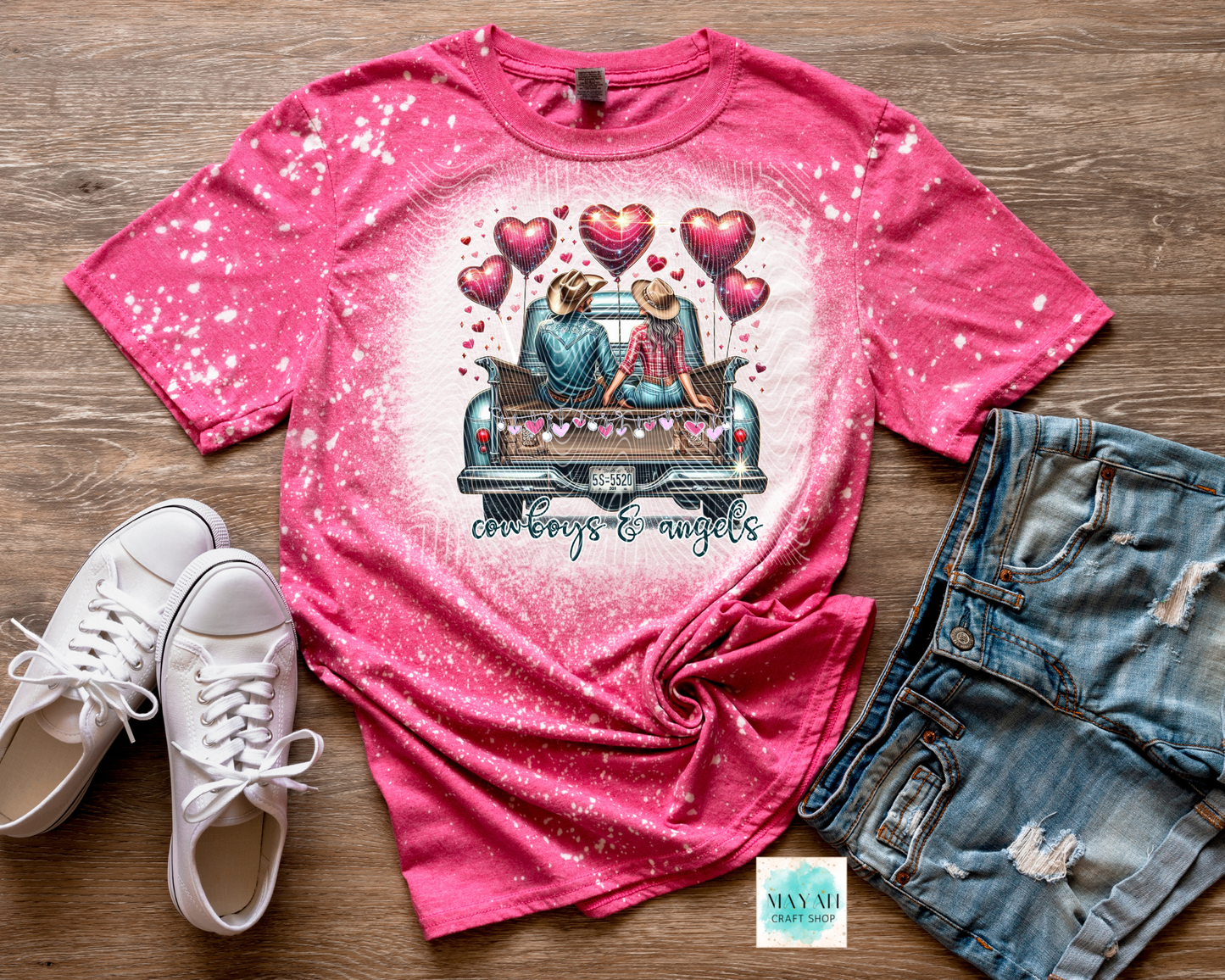 Pink t-shirt with graphic design of two people in a car and hearts, paired with white sneakers and denim shorts on a wooden surface.