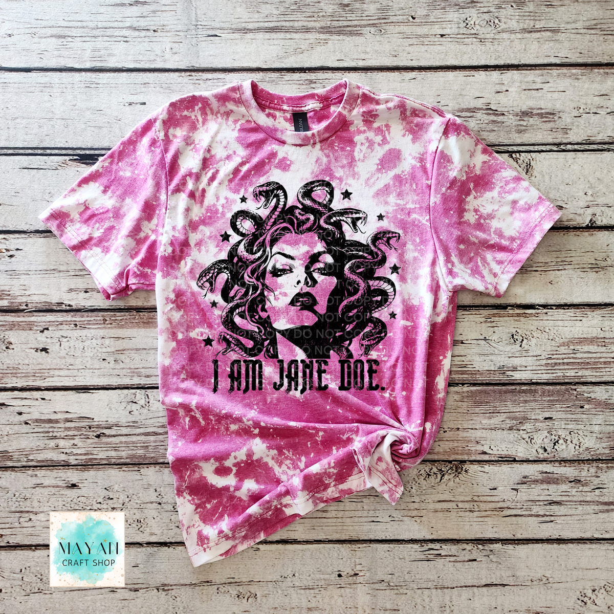 A heather heliconia bleached graphic t-shirt featuring a black illustration of Medusa and the text 'I AM JANE DOE.' laid flat on a rustic wooden plank background.