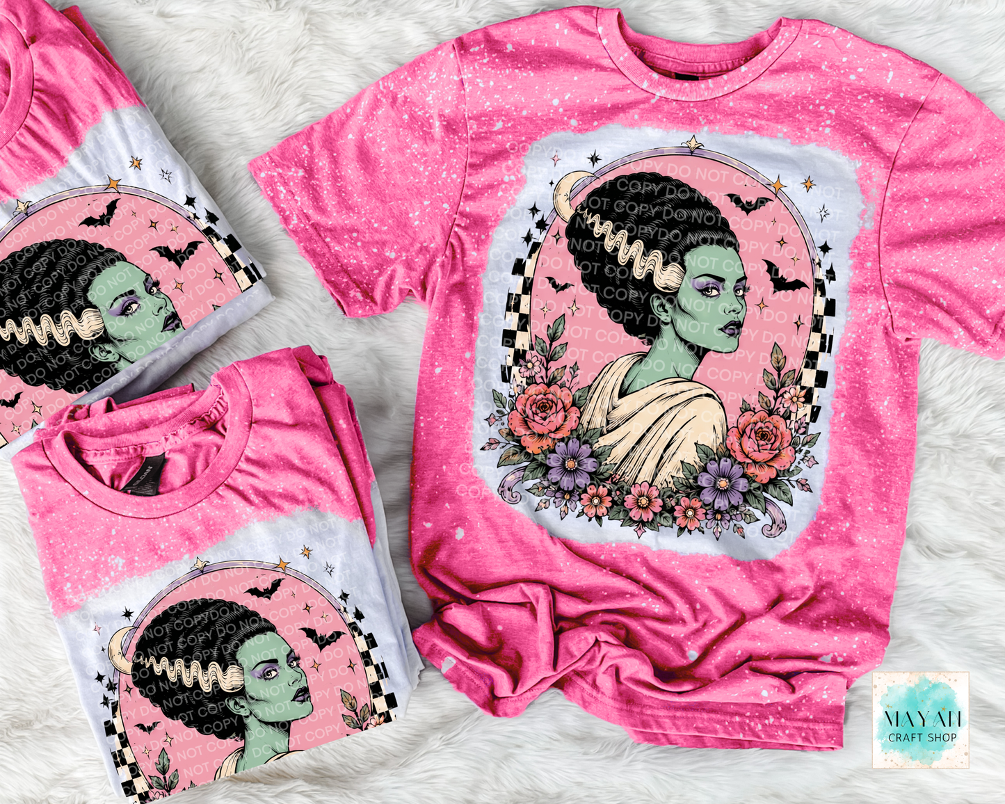 Pink t-shirt with a graphic design of a woman with green skin and black hair, surrounded by flowers and bats, on a textured surface.