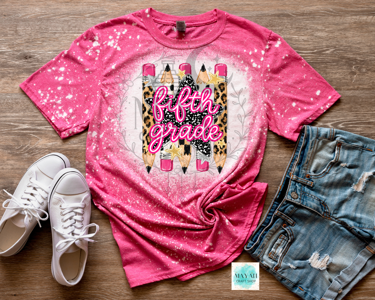 A pink short-sleeve graphic T-shirt laid flat on a wood surface, styled with white sneakers and distressed denim shorts. The shirt has a bright pink splatter/glitter finish and a central graphic showing pencils with leopard-print and black sections, plus the words 'fifth grade' in pink script.