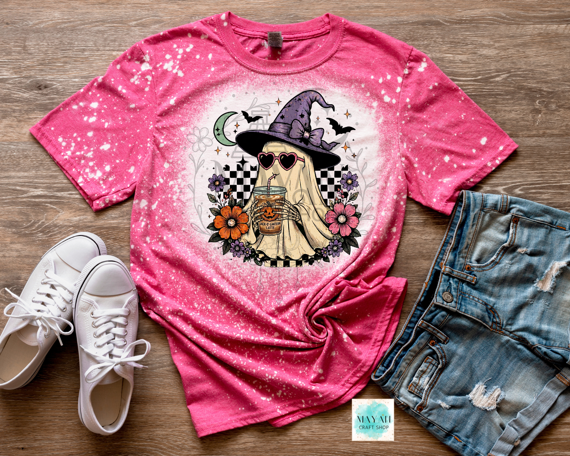 Pink t-shirt with a graphic design of a witch holding a drink, paired with white sneakers and denim shorts on a wooden surface.