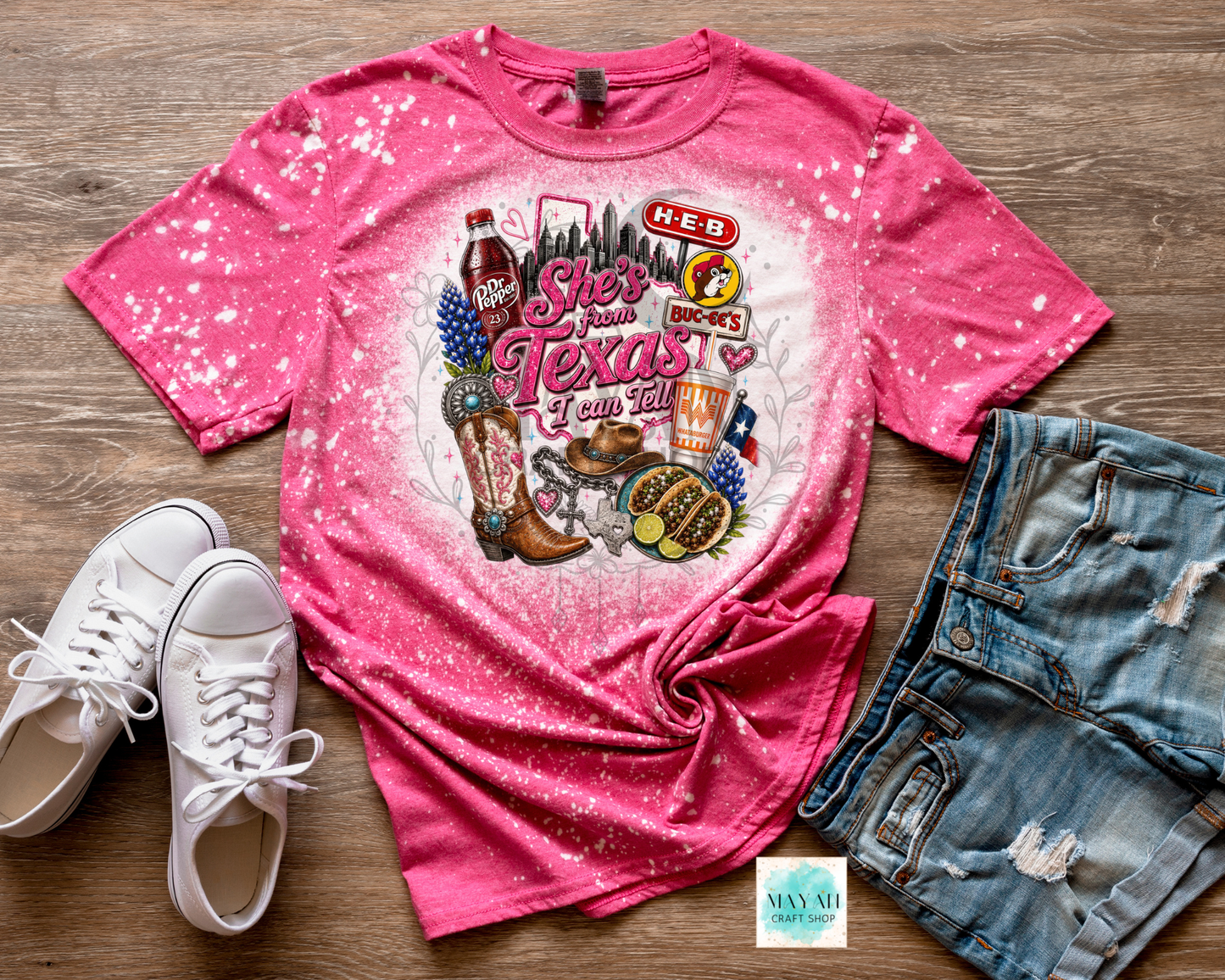 Pink t-shirt with Texas-themed graphics on a wooden surface with white sneakers and denim shorts.
