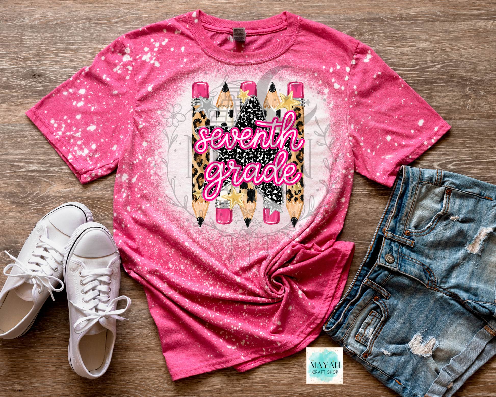 A pink short-sleeve graphic T-shirt laid flat on a wood surface, styled with white sneakers and distressed denim shorts. The shirt has a bright pink to lighter pink gradient with white splatter details and a central graphic of pencils and the words 'seventh grade' in script.