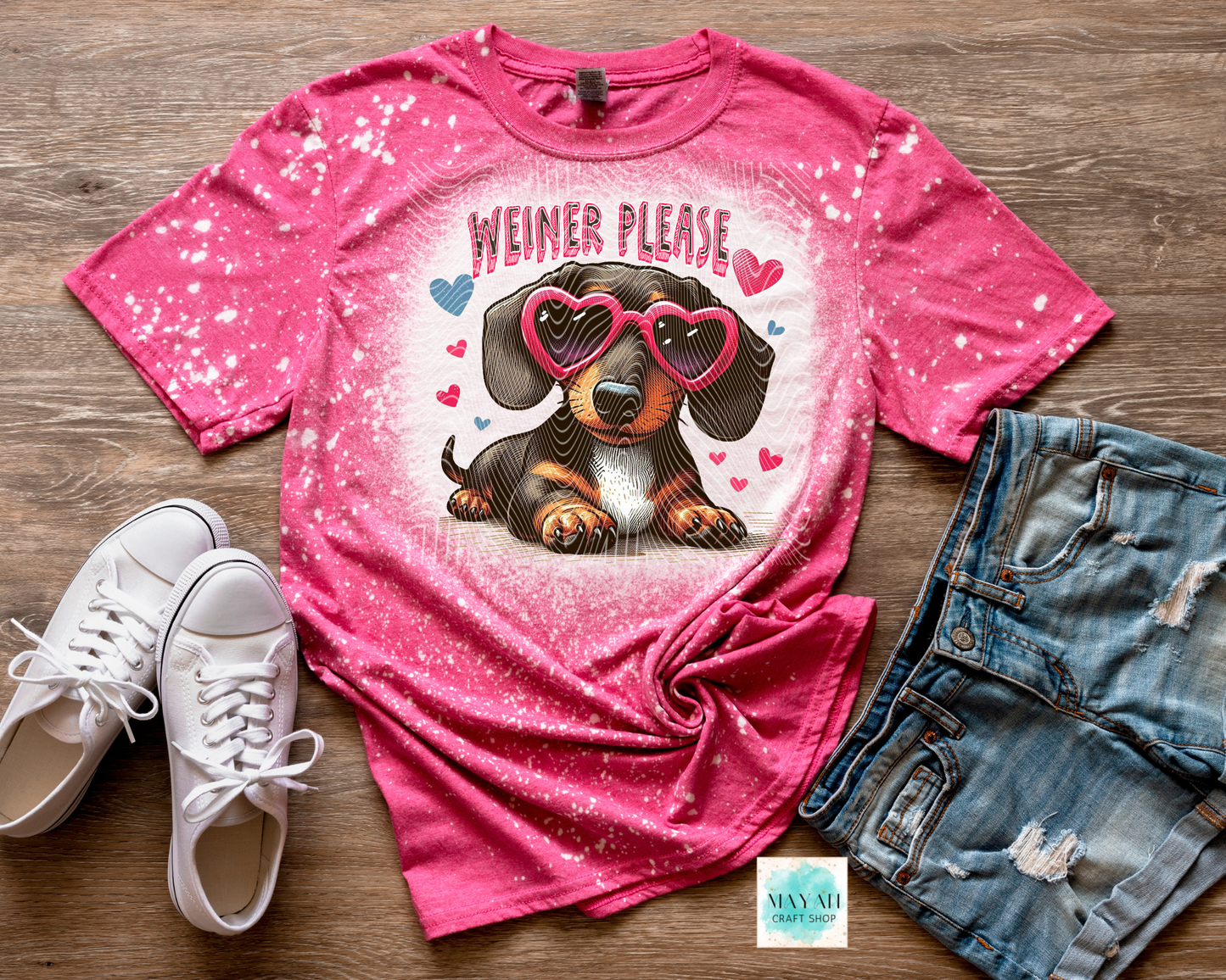 Pink t-shirt with a dachshund graphic and ' Weiner Please' text, paired with white sneakers and denim shorts on a wooden surface.