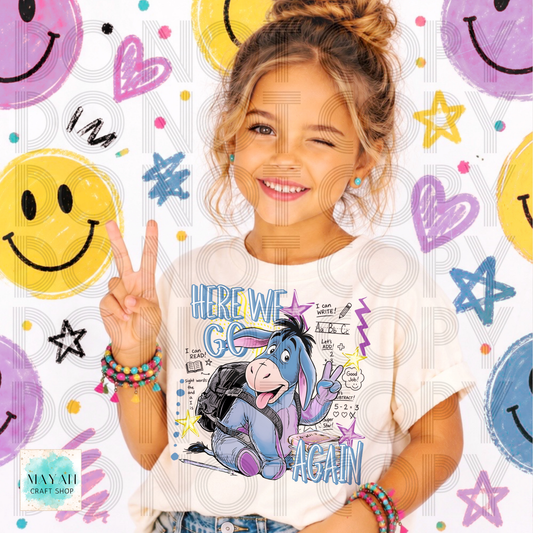 A young girl with her hair in a bun smiles while wearing a white graphic t-shirt featuring a cartoon illustration. She is posing with a peace sign against a white background decorated with colorful faces, hearts, stars, and doodles.