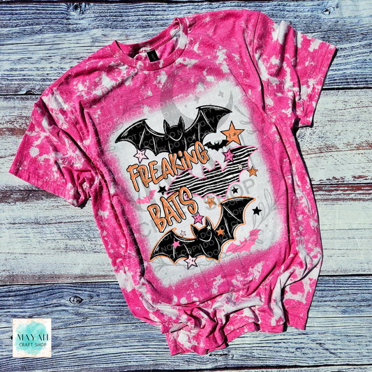 A pink bleached graphic t-shirt featuring two black bats and the words 'FREAKING BATS' in orange, laid flat on a rustic gray wood plank background.