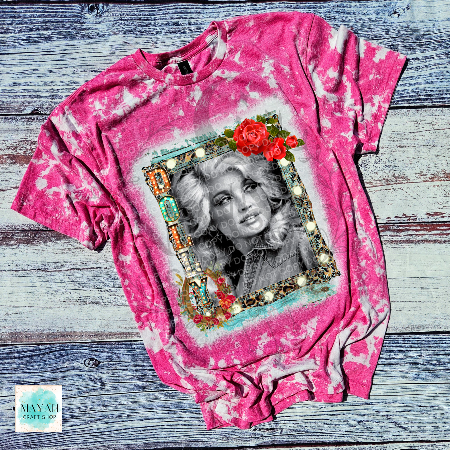 A pink acid-wash graphic t-shirt featuring a black and white portrait of a country singer inside an ornate gold frame with red roses, laid flat on a rustic gray wood plank background.
