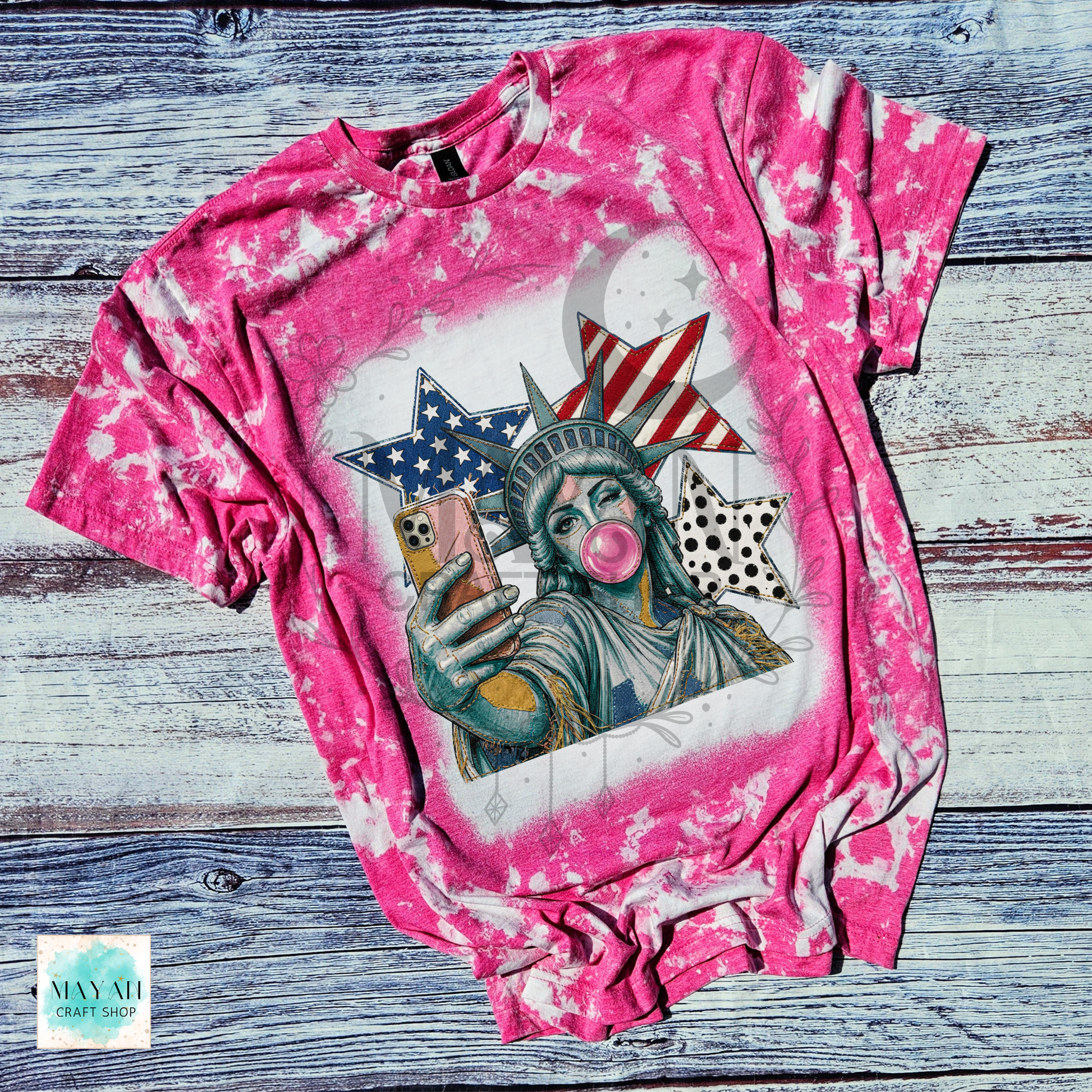 Pink tie-dye t-shirt with a graphic of the Statue of Liberty taking a selfie on a wooden surface.