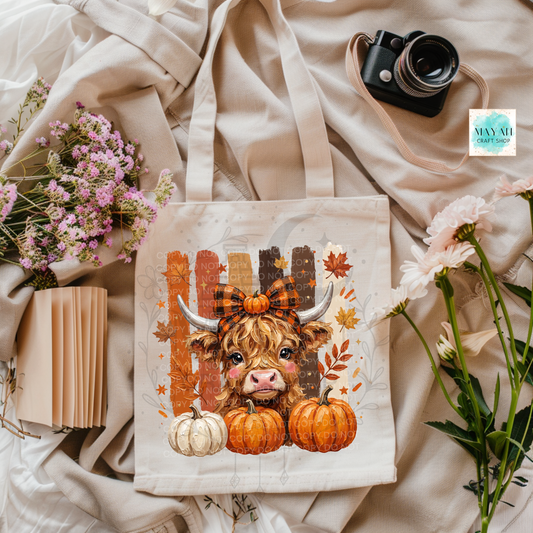 A canvas tote bag with a fall-themed Highland cow illustration, styled in a flat lay with beige fabric, flowers, a camera, and a notebook.