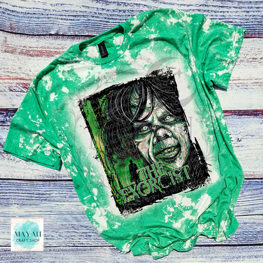 A green bleached graphic t-shirt featuring a stylized portrait of a character with a text on the front, laid flat on a rustic whitewashed wooden background.