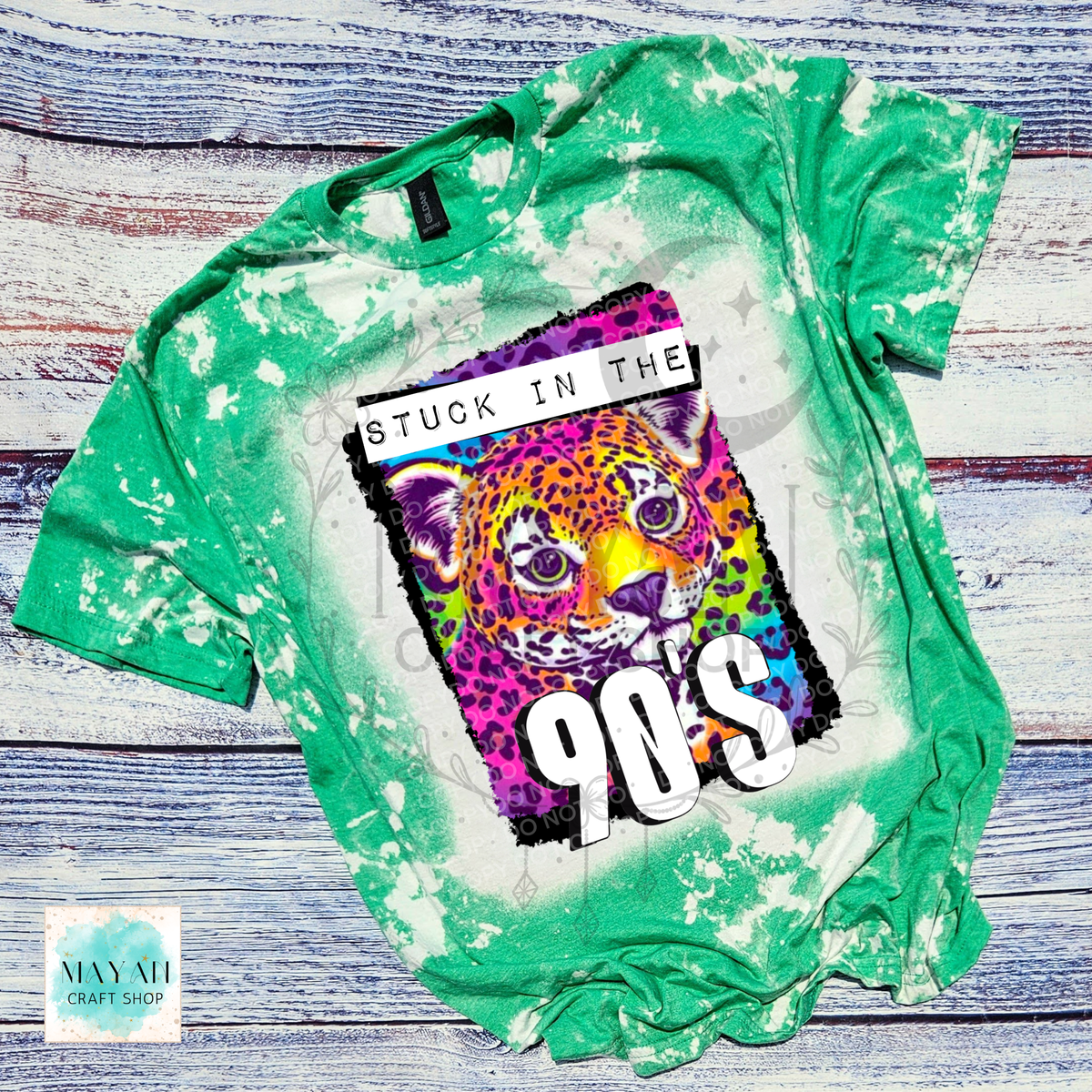 A green bleached t-shirt with a colorful graphic of a leopard and the text 'STUCK IN THE 90S', laid flat on a rustic blue wood background.