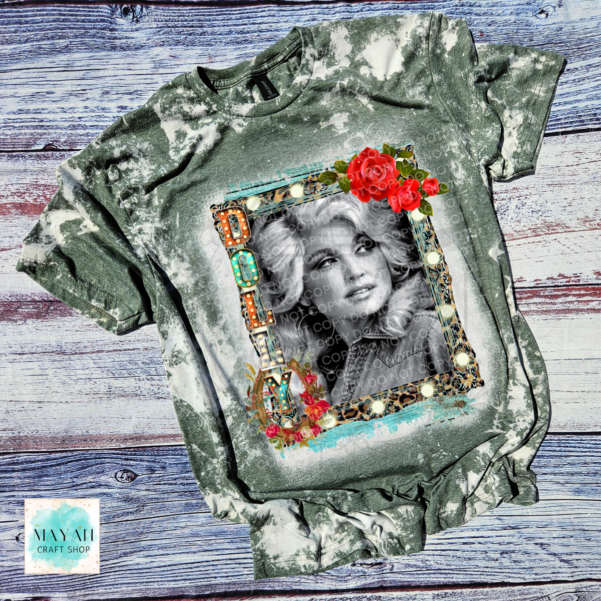 A short-sleeve graphic t-shirt with a green and white bleach tie-dye pattern, featuring a central portrait of a country singer inside a leopard-print frame with red roses, laid flat on a rustic blue wood background.