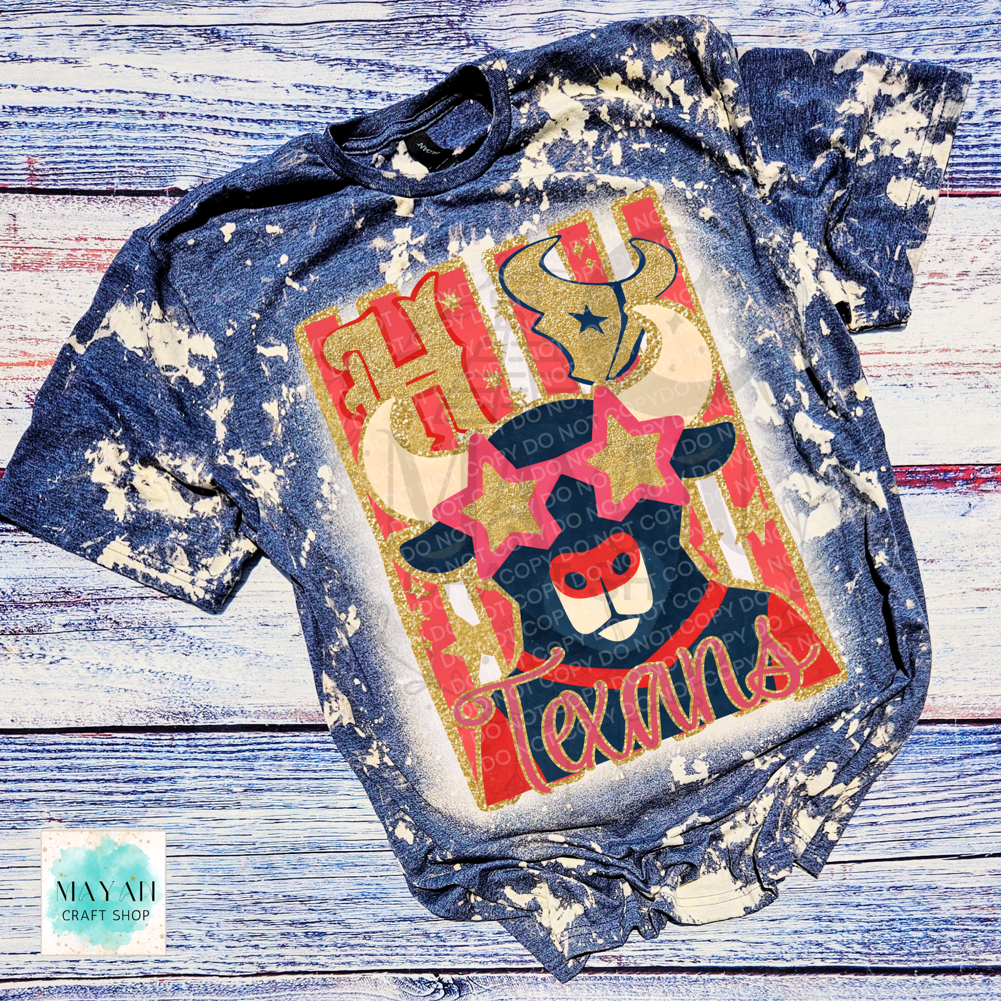 A short-sleeve graphic T-shirt laid flat on a rustic whitewashed wood surface. The shirt has bleach-splatter finish and features a large front graphic with words in red and gold, a bull, a bear, and red-and-gold stars.