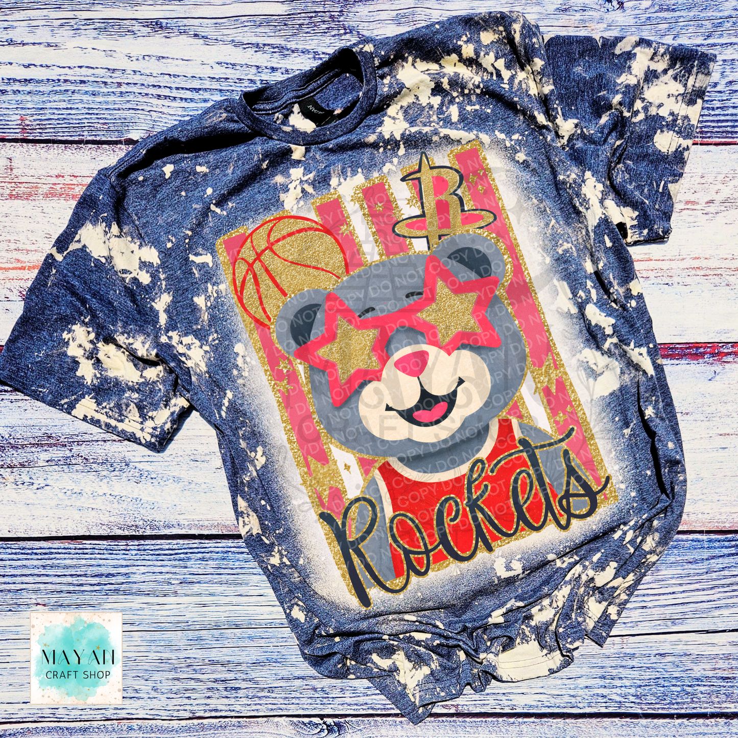 A short-sleeve graphic T-shirt laid flat on a rustic whitewashed wood background. The shirt has a distressed bleach-splatter finish and features a large front graphic of a gray bear wearing red star-shaped sunglasses, with a word and basketball-themed graphics.