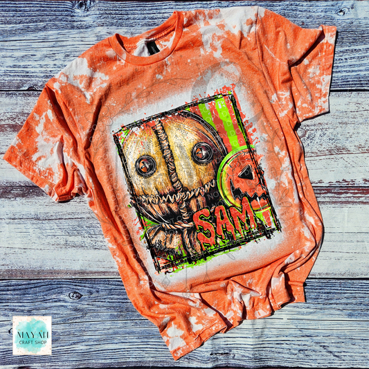An orange bleached graphic t-shirt featuring a horror character design with a text on the front, laid flat on a rustic gray wood plank background.