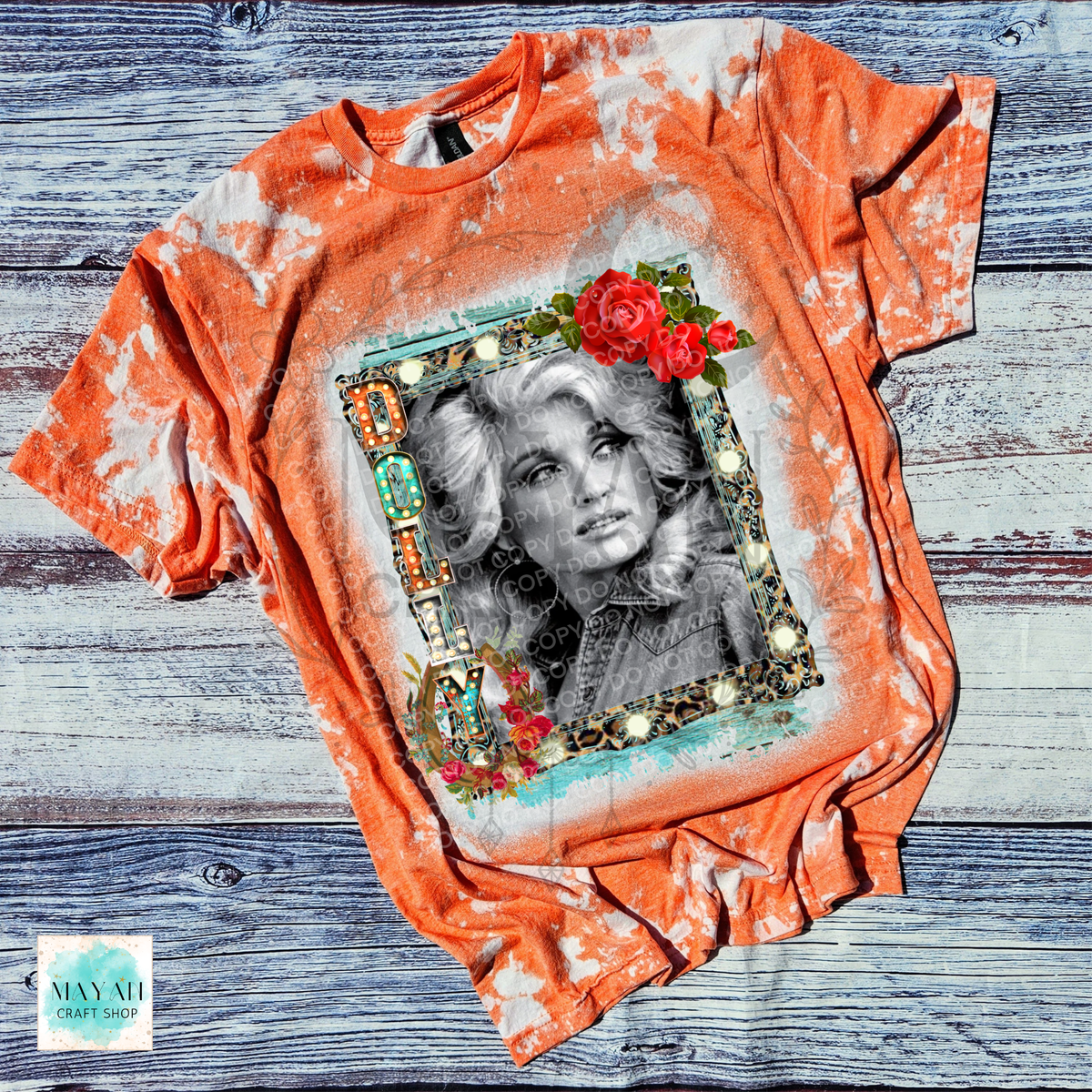 An orange and white bleach-dyed graphic t-shirt featuring a black and white portrait of a country singer inside a decorative frame with red roses, laid flat on a rustic gray wood background.