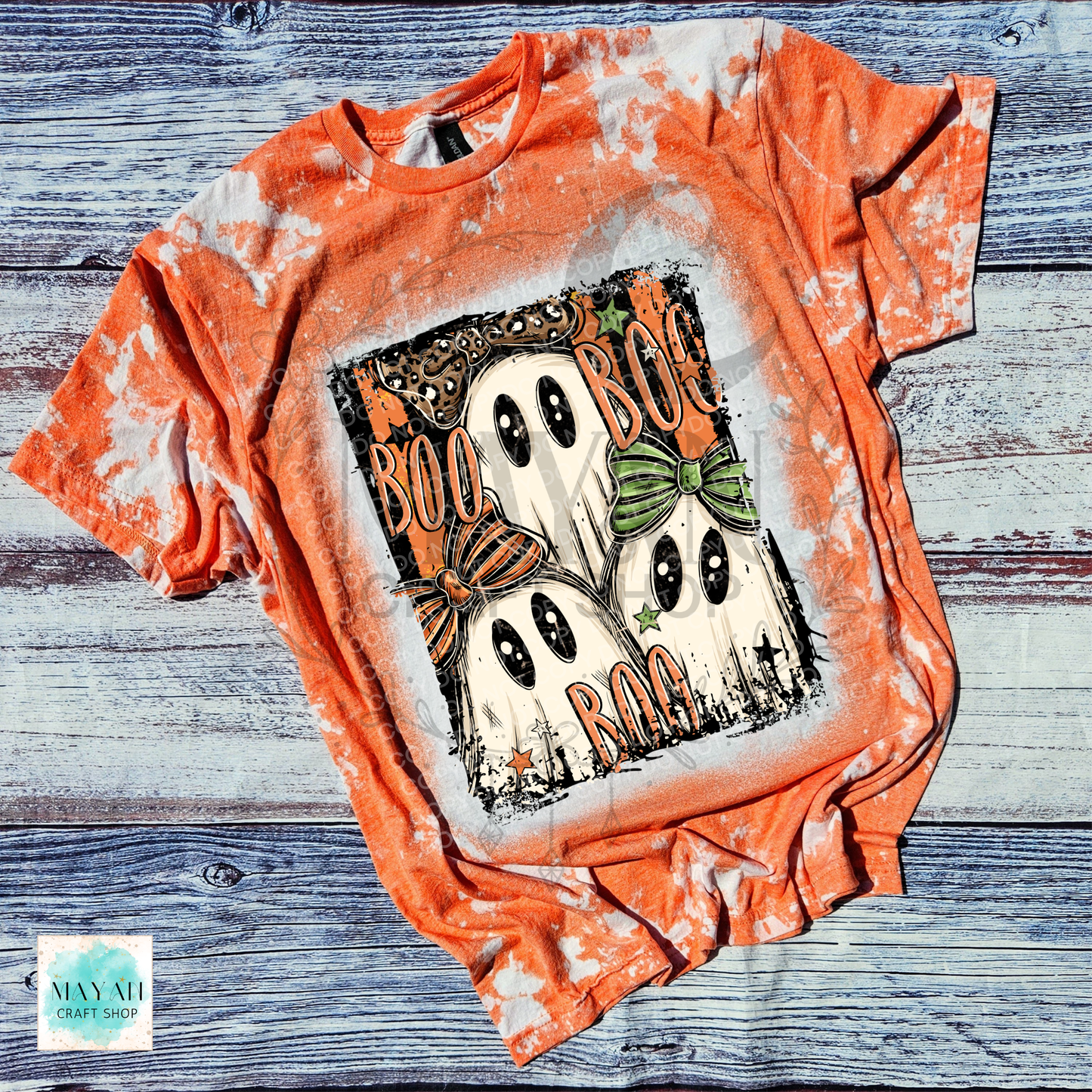 An orange bleached t-shirt featuring a Halloween graphic of two ghosts with bows and the text 'BOO BOO BOO'. The shirt is laid flat on a rustic blue-gray wooden background.