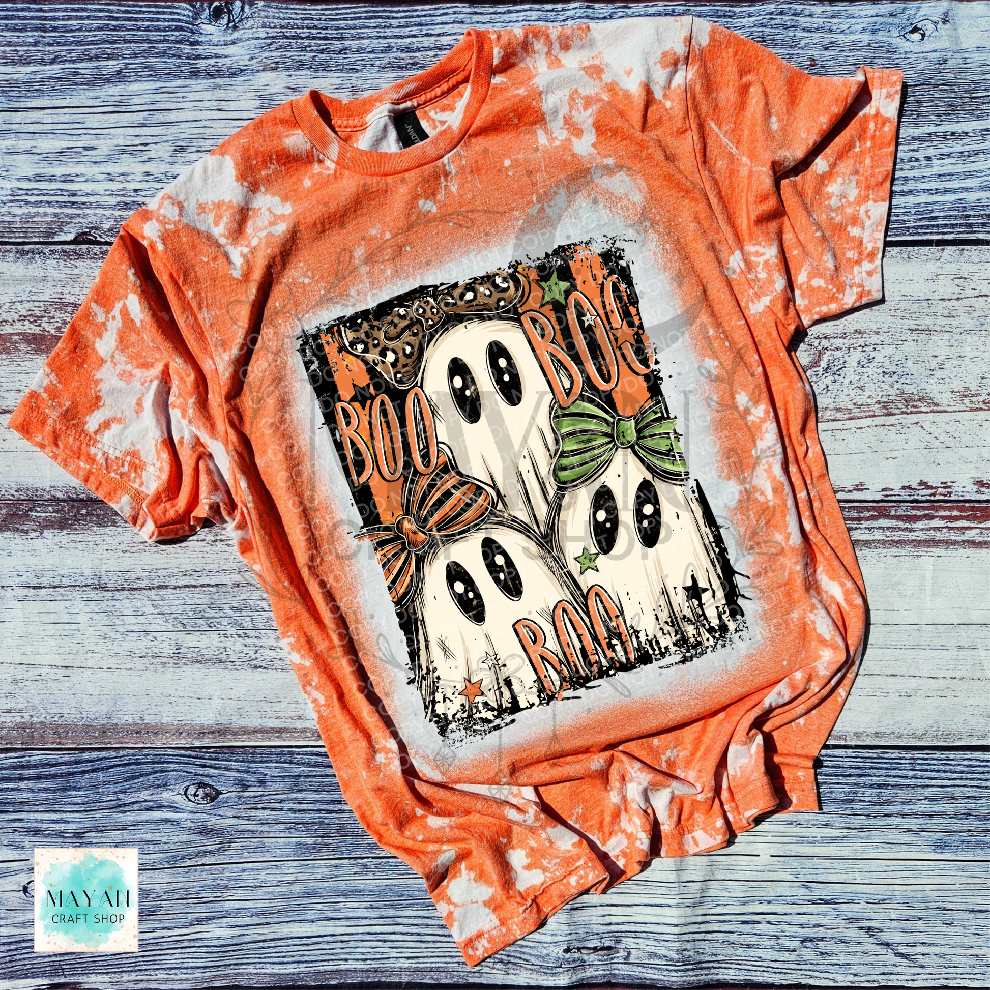 An orange bleached t-shirt featuring a Halloween graphic of two ghosts with bows and the text 'BOO BOO BOO'. The shirt is laid flat on a rustic blue-gray wooden background.