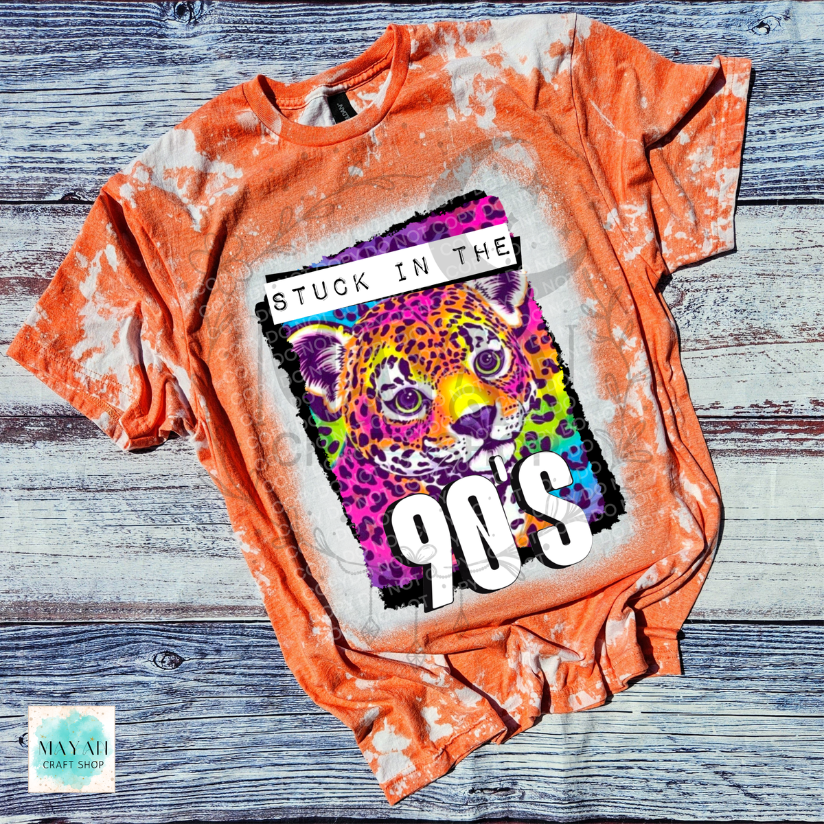 An orange bleached graphic t-shirt laid flat on a rustic gray wood background. The graphic features a colorful leopard illustration with the text 'STUCK IN THE 90's'.