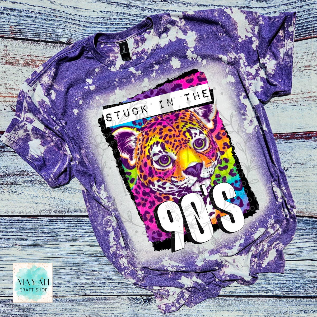 A purple bleached graphic t-shirt laid flat on a rustic blue-gray wooden background. The shirt features a large graphic print of a colorful leopard with the text 'STUCK IN THE 90's'.