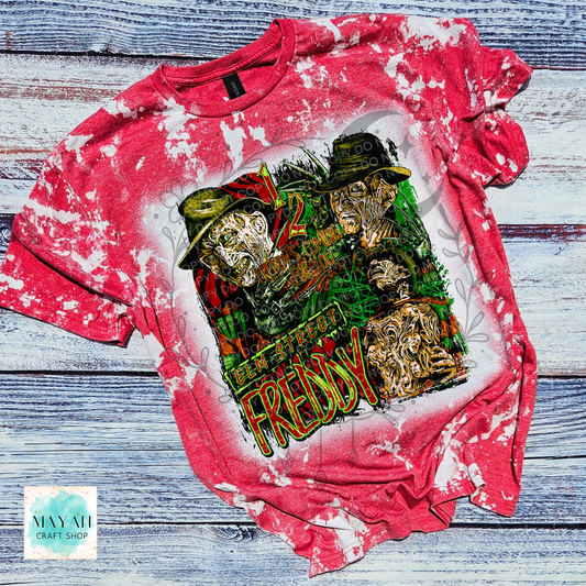 A red bleached graphic t-shirt featuring a horror-themed collage of a horror character with a text. The shirt is laid flat on a rustic, weathered blue-gray wooden plank background.