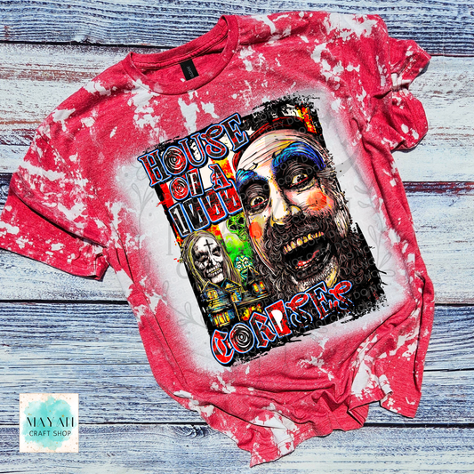 A red and white bleached graphic t-shirt laid flat on a rustic blue-gray wooden background. The shirt features a large, colorful graphic print of a creepy clown face with a text.