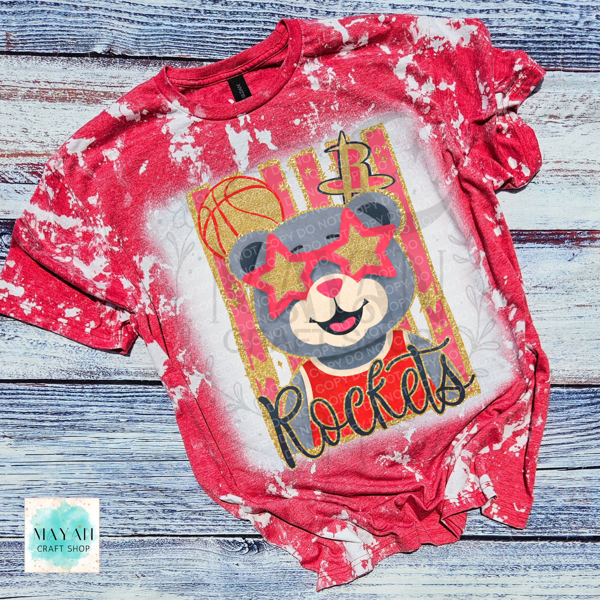 A red and white bleach-dyed graphic t-shirt featuring a cartoon bear wearing star-shaped sunglasses with a basketball team logo and a text, laid flat on a rustic blue-gray wooden background.