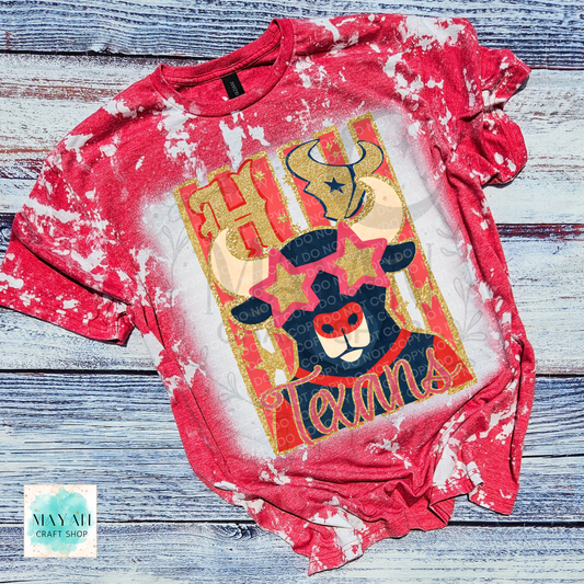 A red and white bleached t-shirt with a bull graphic on the front, laid flat on a rustic blue-gray wooden background. The graphic features a bull wearing a cowboy hat with stars and a word in script.