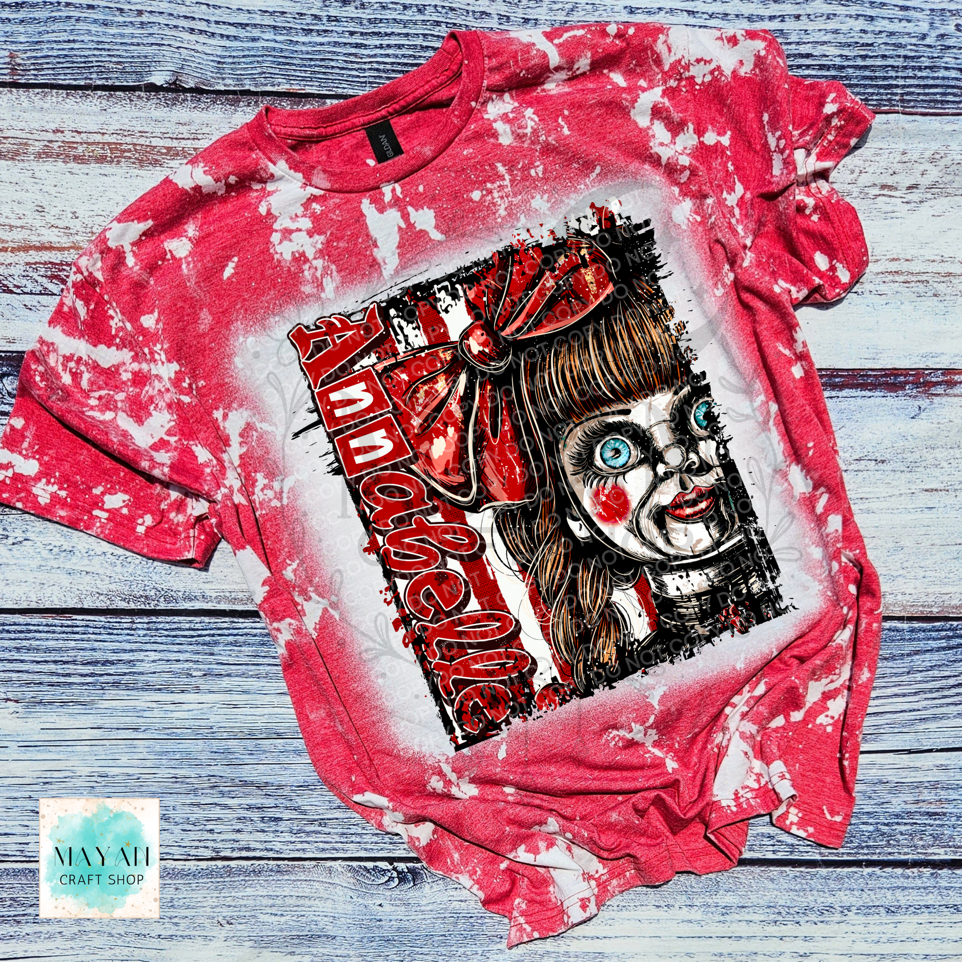 A red and white bleached graphic t-shirt featuring a horror-style illustration of a creepy doll character with a text on the front, laid flat on a rustic blue-gray wooden background.