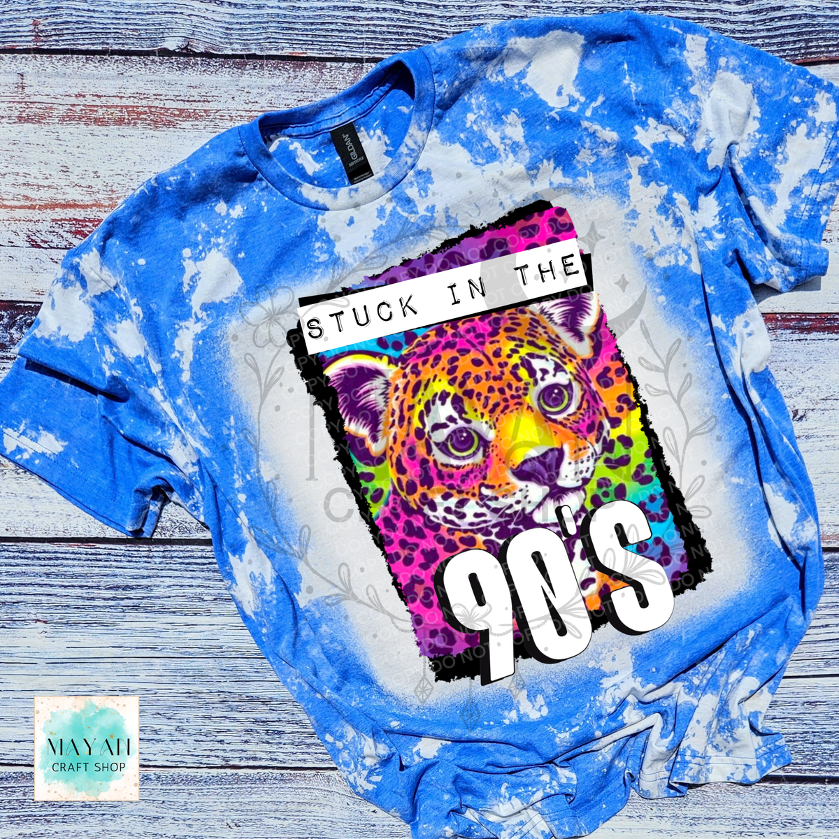 A blue bleached t-shirt featuring a colorful graphic of a leopard with the text 'STUCK IN THE 90'S'. The t-shirt is laid flat on a rustic wooden background.