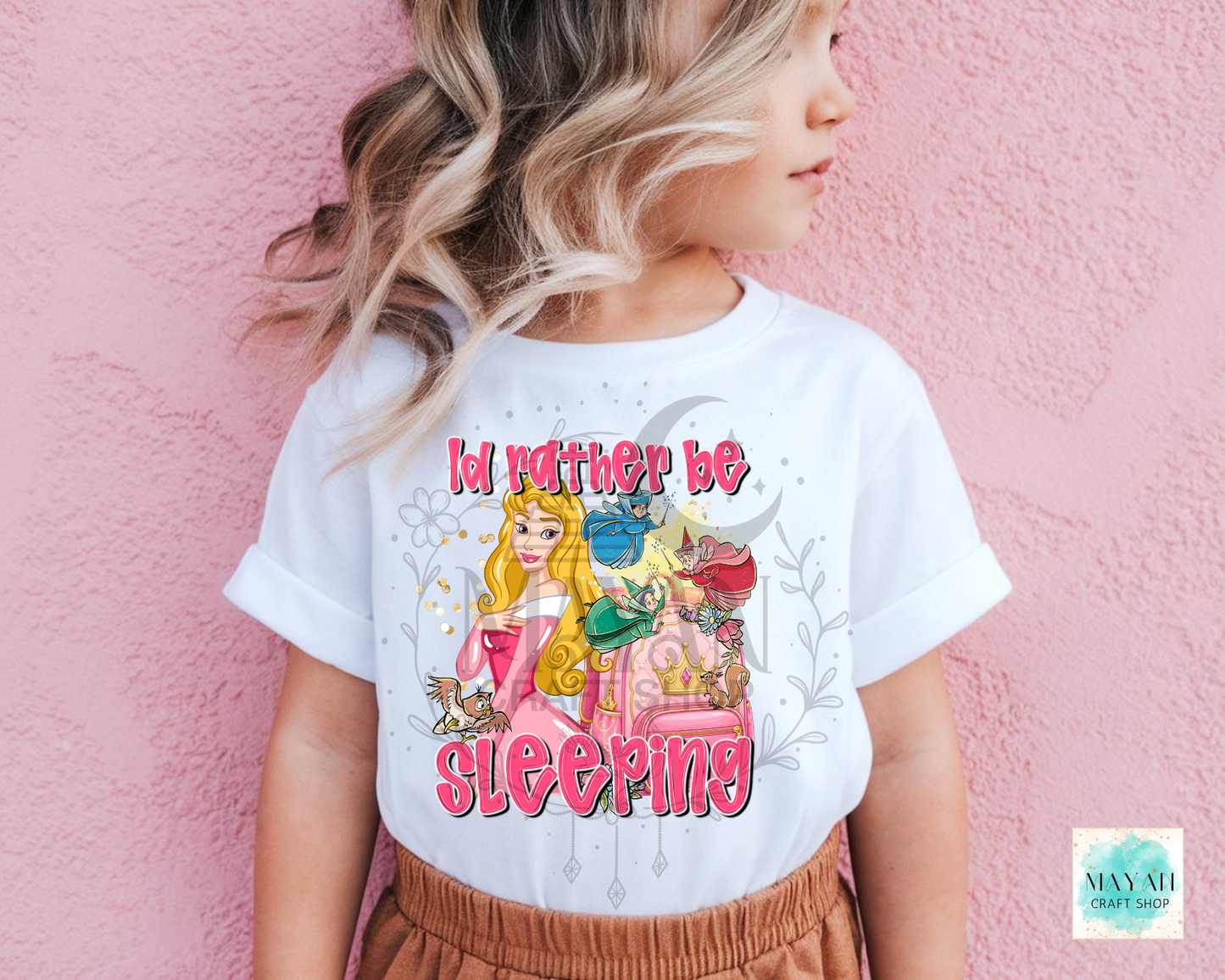A child wearing a white graphic T-shirt with a colorful illustration of a princess and the text 'I'd rather be sleeping', posed against a pink textured wall.