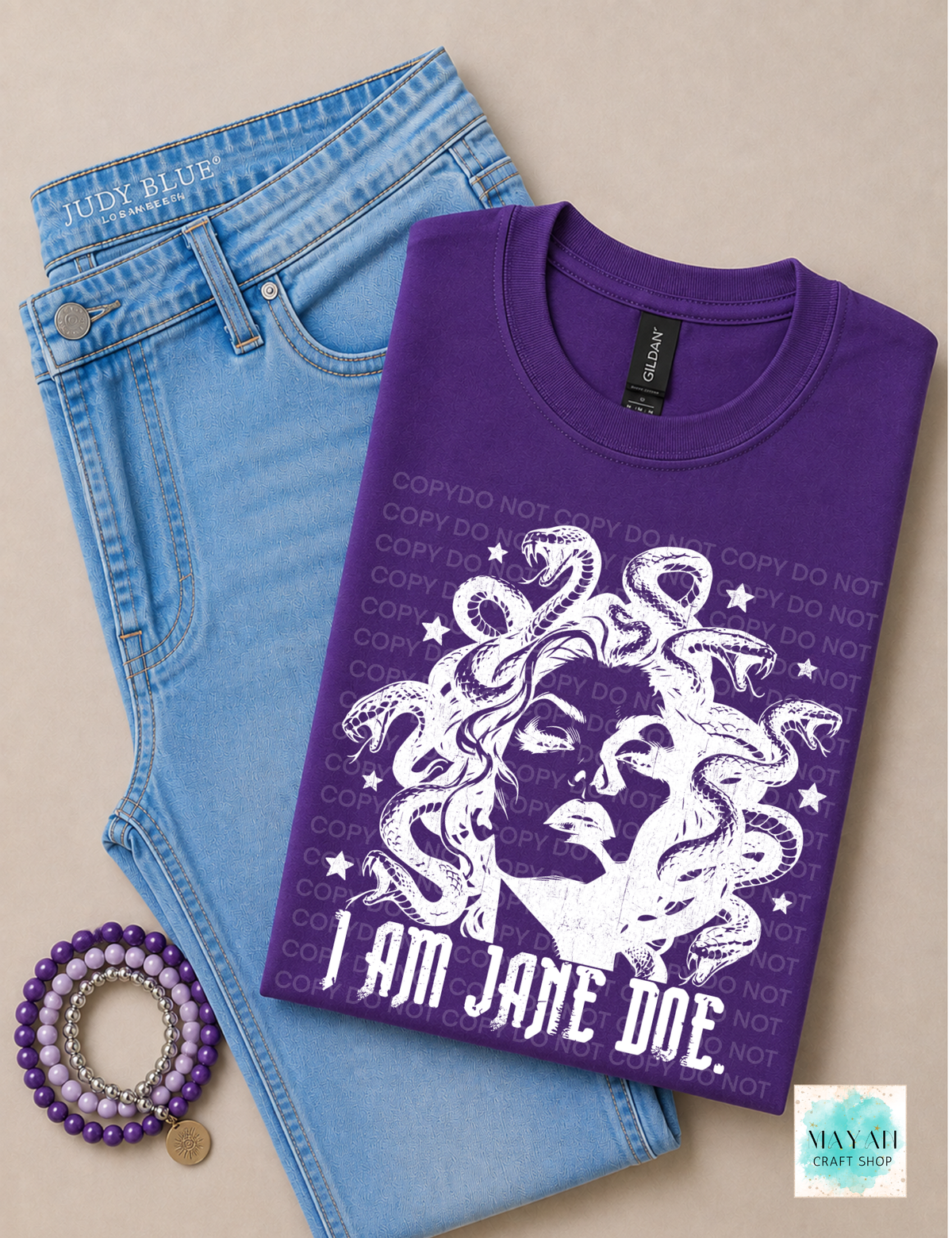 A folded purple graphic T-shirt with a white illustration and the text 'I AM JANE DOE.' is styled on top of light blue jeans on a beige background, with a purple beaded bracelet nearby.