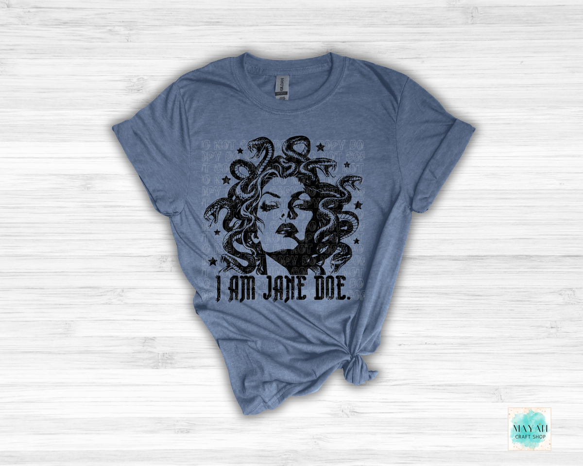 A heathered indigo blue short-sleeve t-shirt with a distressed black graphic of Medusa and the text 'I AM JANE DOE.' displayed on a white wood-plank background.