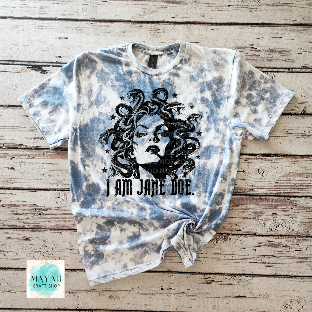 A short-sleeve graphic T-shirt laid flat on a rustic wood plank background. The shirt is heather indigo blue bleached, with a large black illustration of Medusa's face and hair and the text 'I AM JANE DOE.