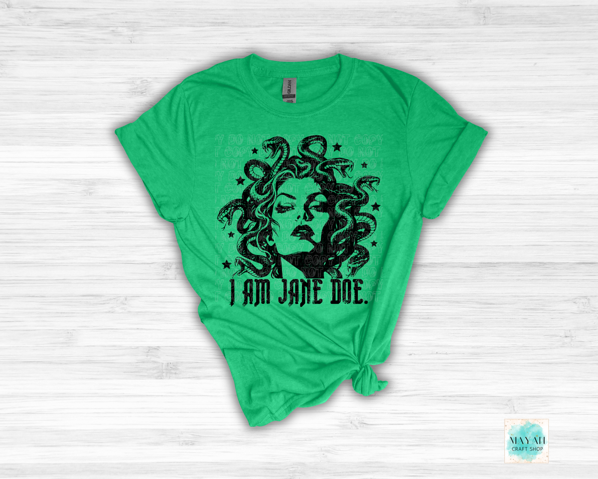 A heather irish green graphic t-shirt featuring a black illustration of Medusa with snakes and the text 'I AM JANE DOE.' printed on the front, styled flat on a white wood background.