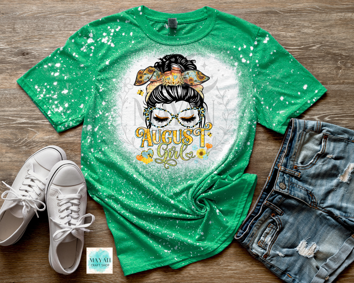A green short-sleeve graphic T-shirt laid flat on a wood surface, styled with white sneakers, distressed denim shorts. The shirt has a white circular glow and paint-splatter effect, featuring a messy bun girl illustration with the words 'August 1st'.
