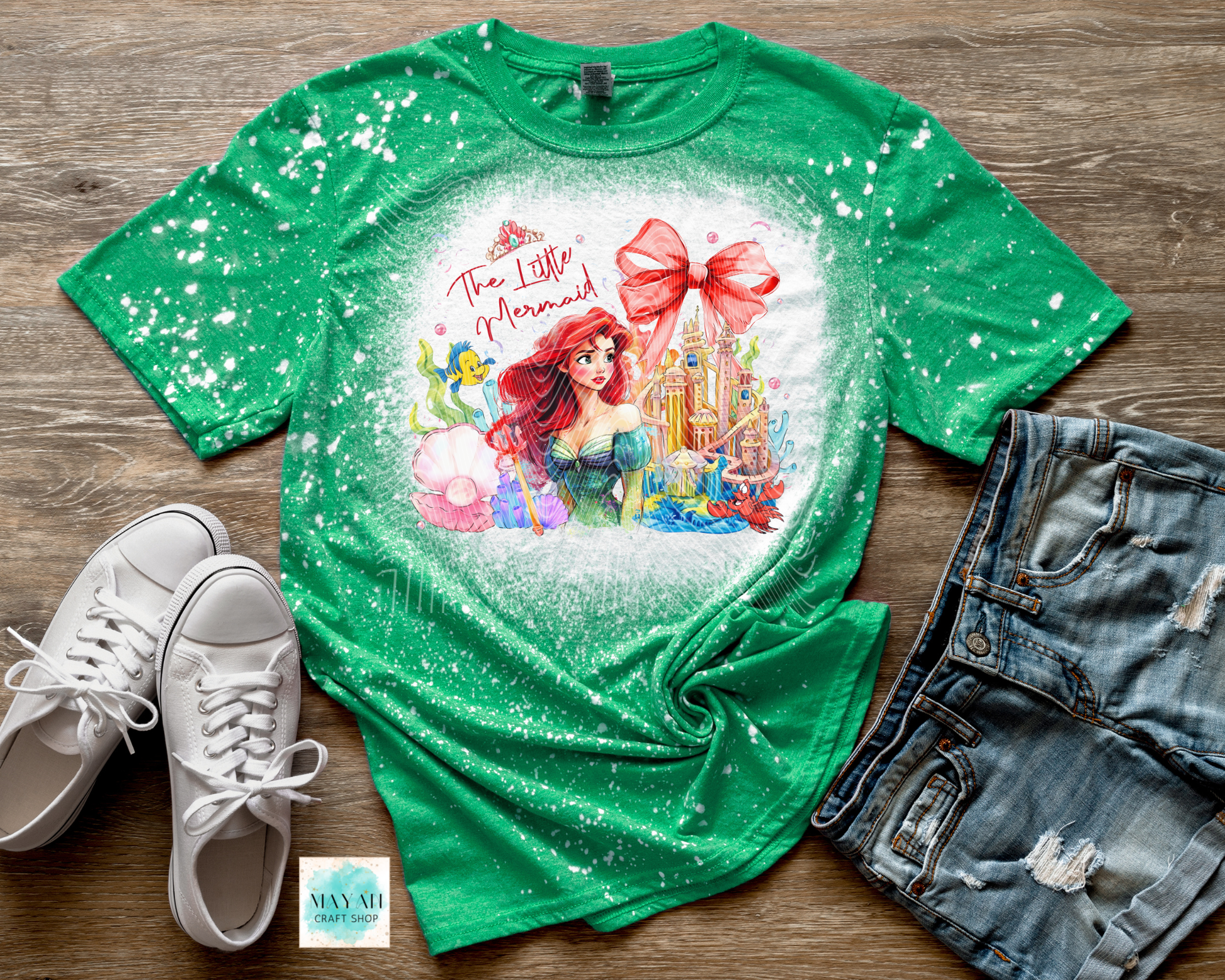 Green t-shirt with a mermaid design on a wooden surface with sneakers and shorts.