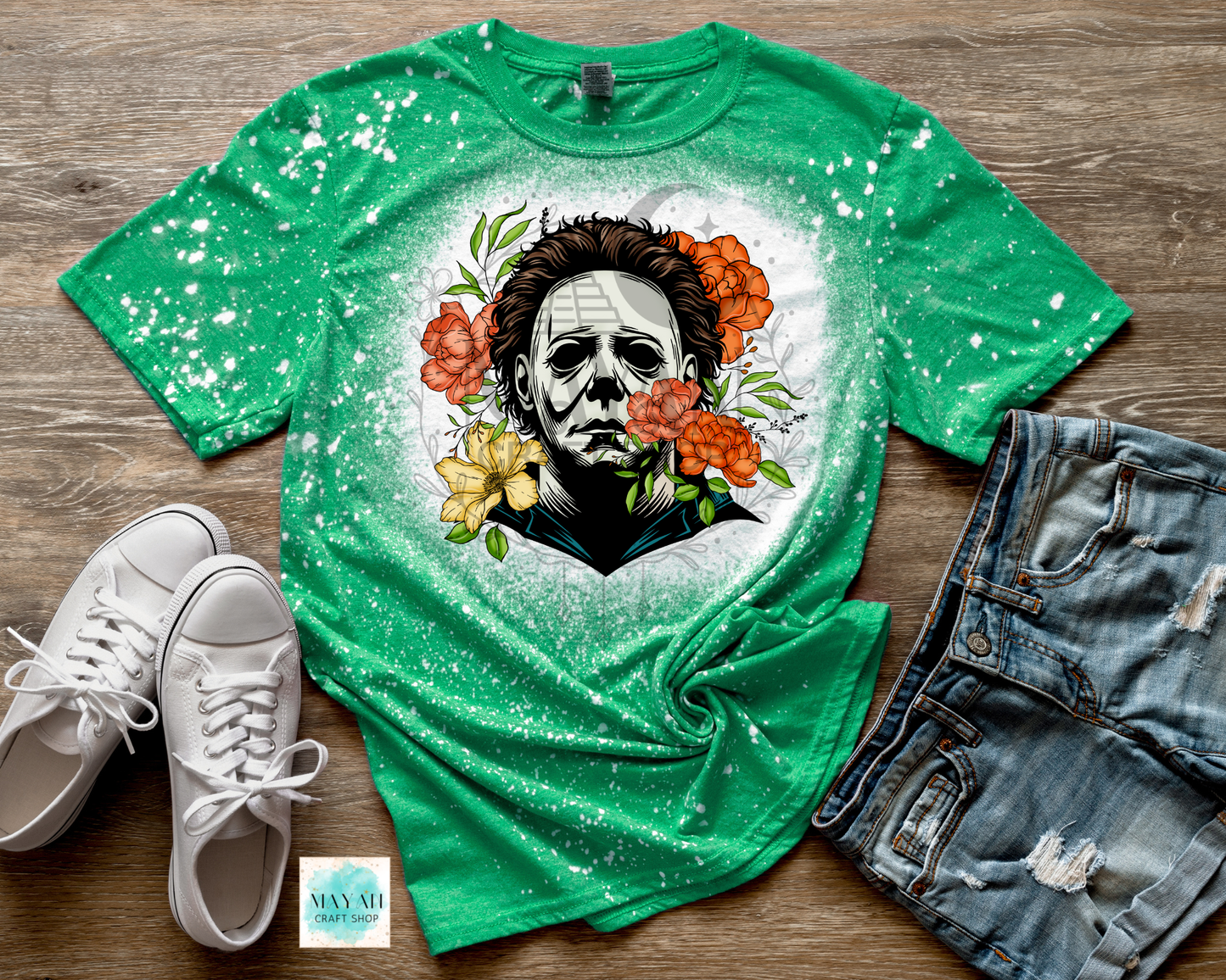 Green t-shirt with a graphic design of a face surrounded by flowers, paired with white sneakers and denim shorts on a wooden surface.