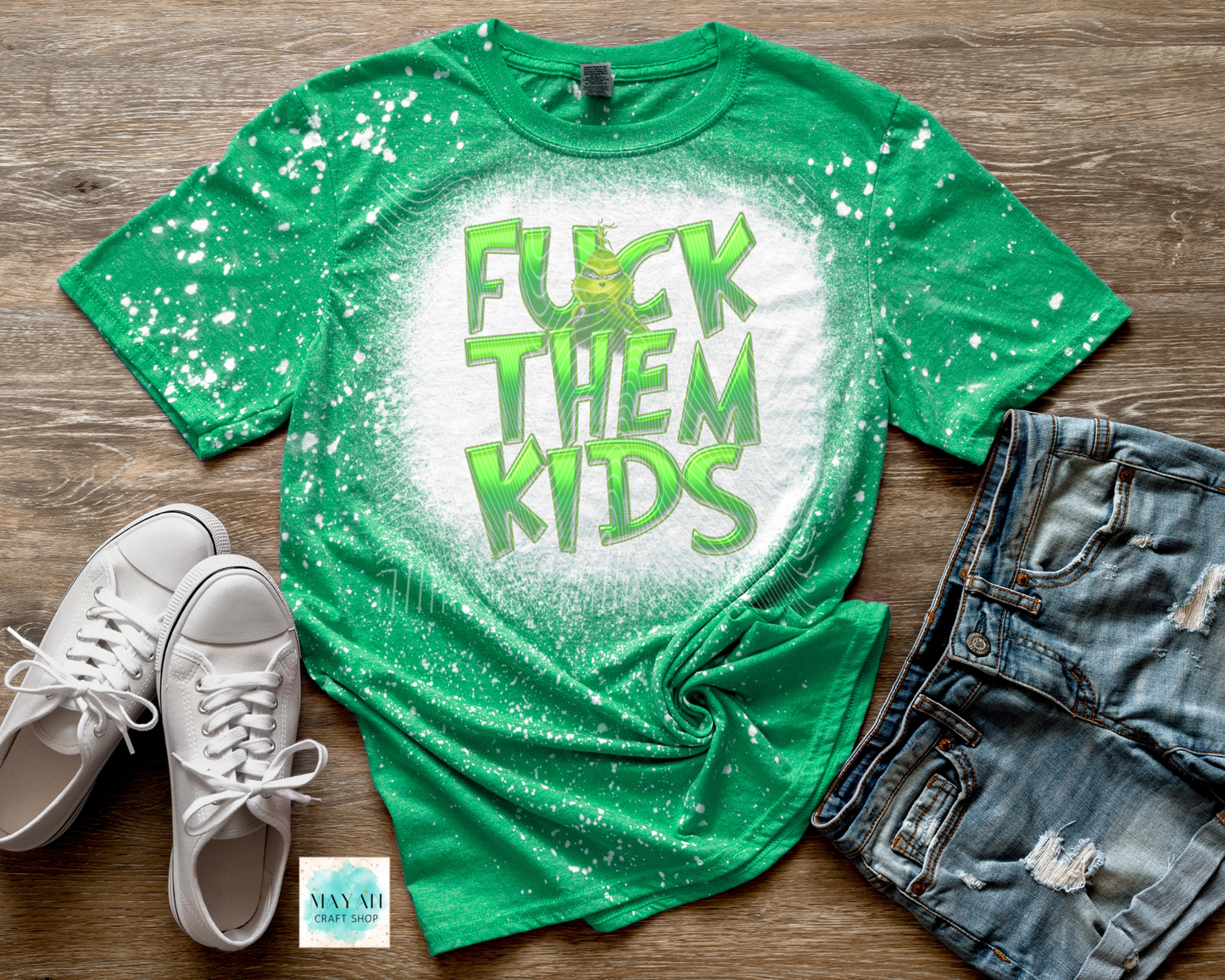 Green t-shirt with 'F**K THEM KIDS' text, white sneakers, and denim shorts on a wooden surface.
