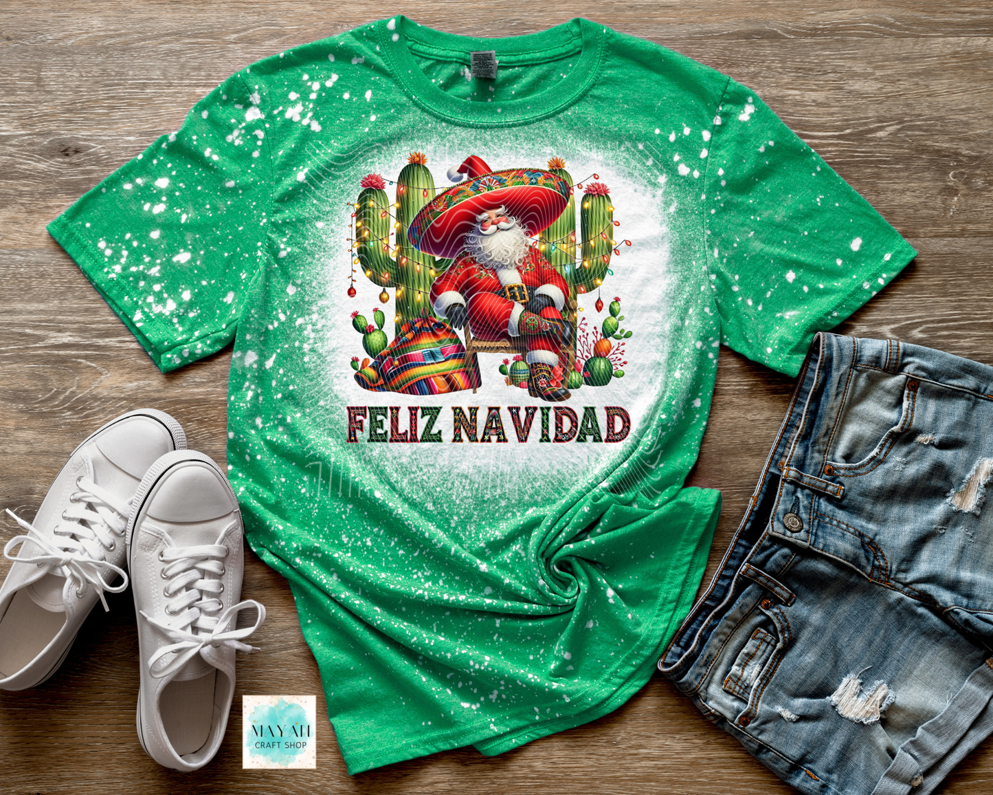 Green t-shirt with Santa Claus and cactus design, text 'Feliz Navidad', white sneakers, and denim shorts on a wooden surface.