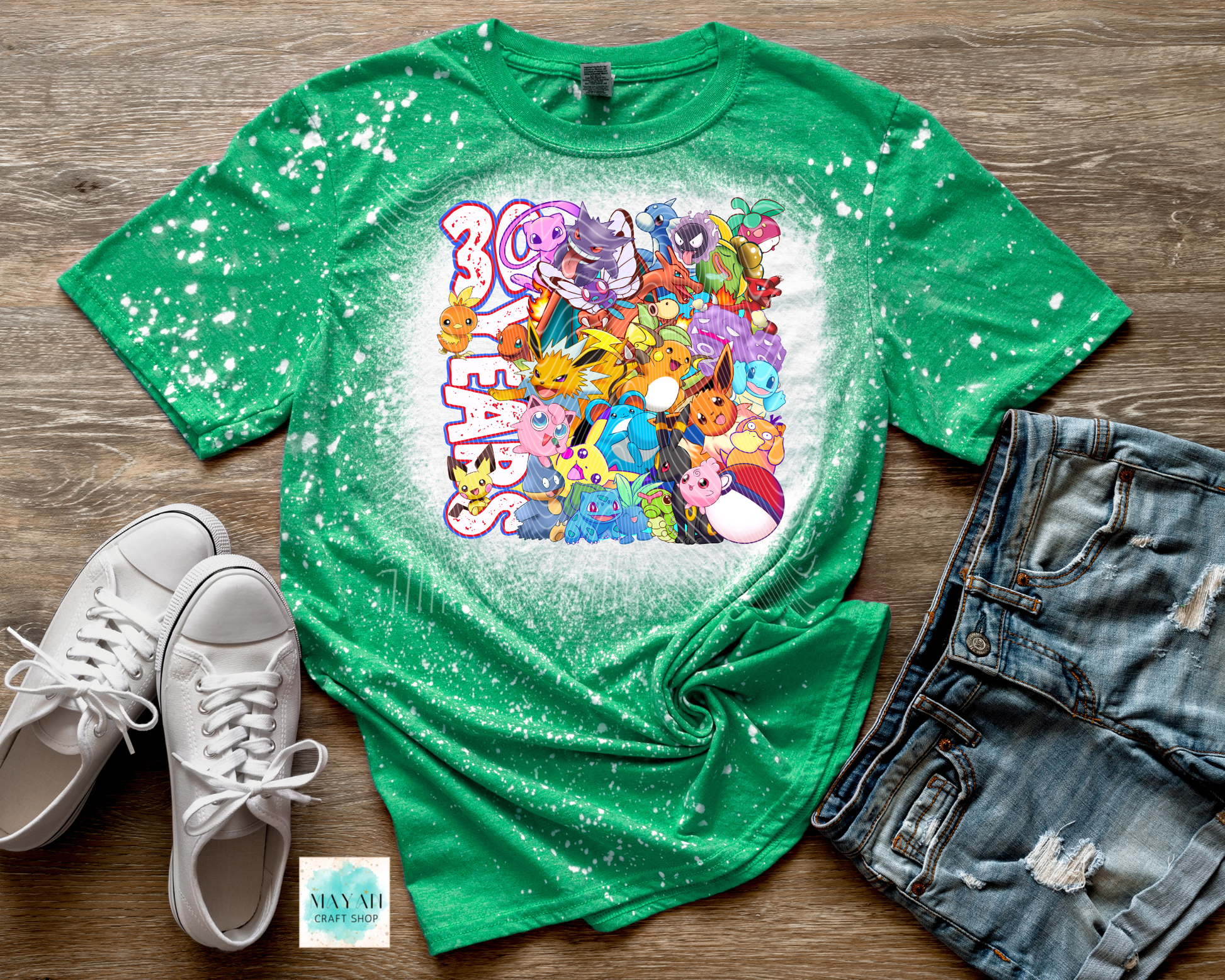 Green t-shirt with colorful graphic design on a wooden surface with white sneakers and denim shorts.