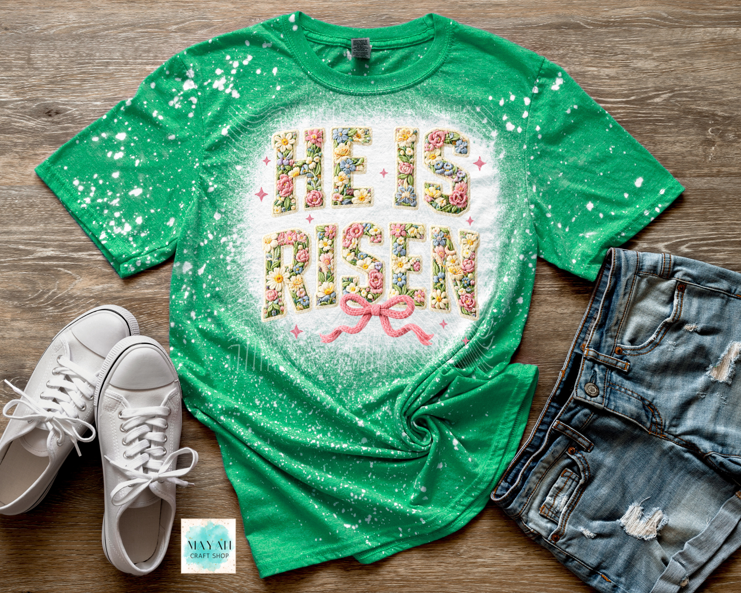 Green t-shirt with 'He is Risen' text, white sneakers, and denim shorts on a wooden surface.